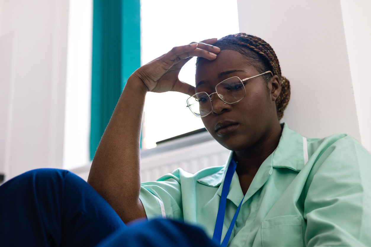 Healthcare professional sitting on the floor with hand on forehead, appearing tired and overwhelmed
