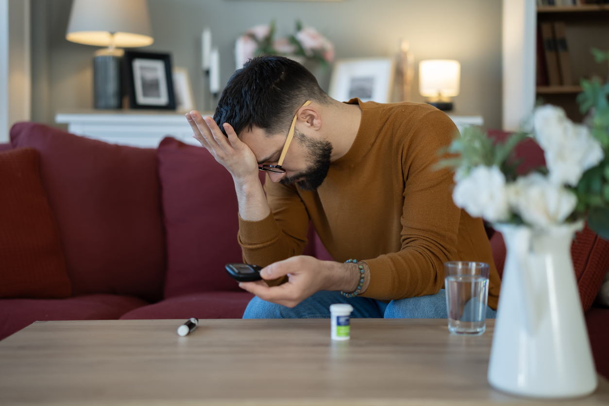 Man sitting at home looking distressed while checking a glucose monitor, illustrating the emotional impact of managing diabetes and blood sugar levels.