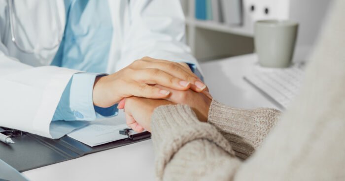 A doctor in a white coat with a stethoscope gently placing their hand on a patient's hand for comfort and reassurance during a medical consultation, with a clipboard and paperwork on the desk