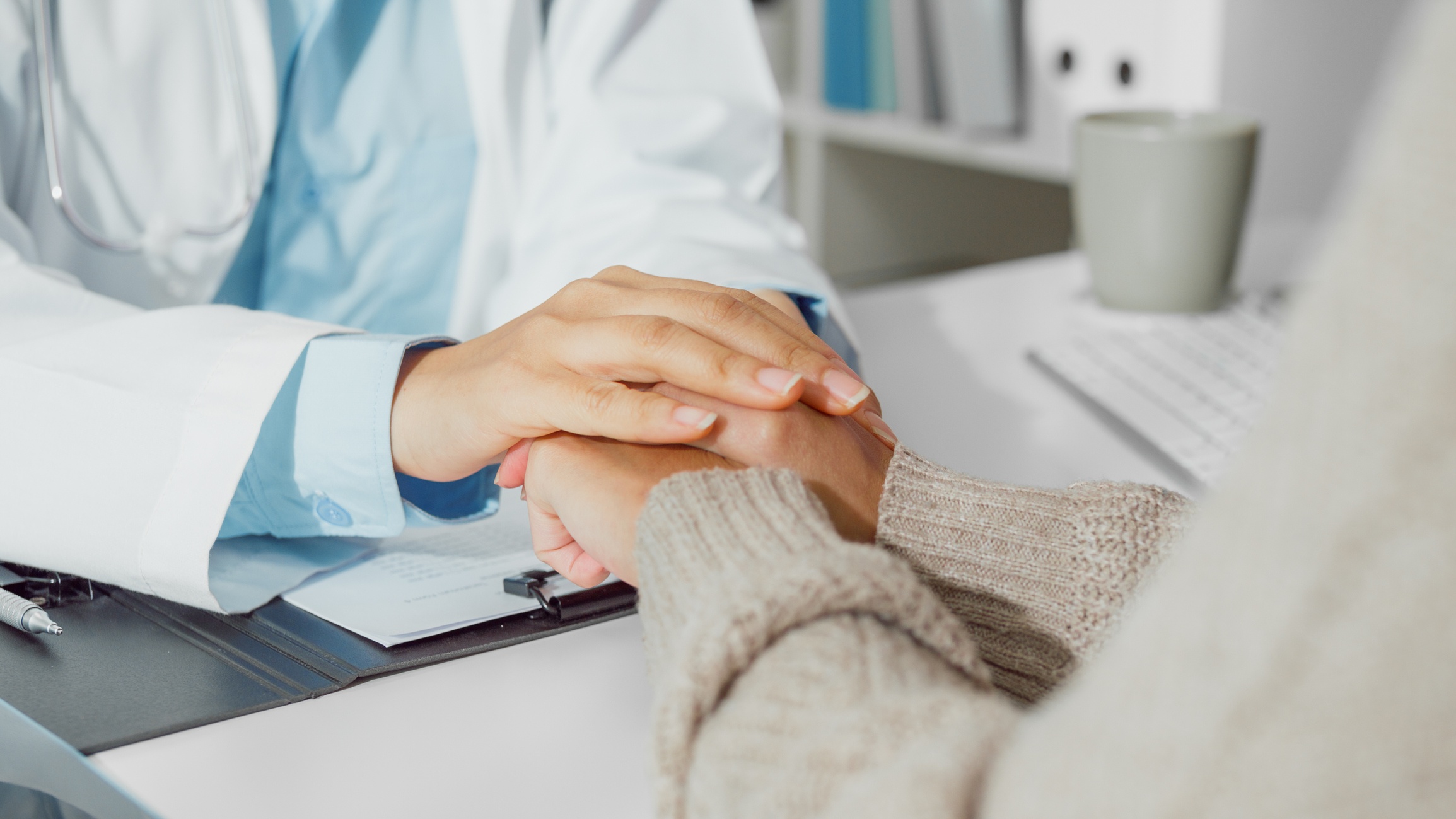 A doctor in a white coat with a stethoscope gently placing their hand on a patient's hand for comfort and reassurance during a medical consultation, with a clipboard and paperwork on the desk