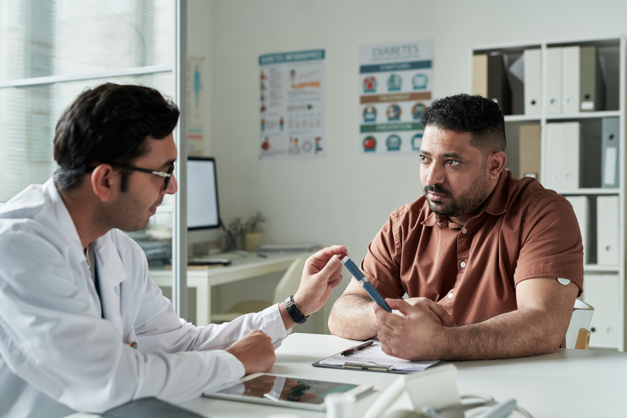 Middle aged Caucasian man sitting at desk consulting with male doctor in medical office, discussing diabetes management while holding glucose meter, diabetes posters on wall