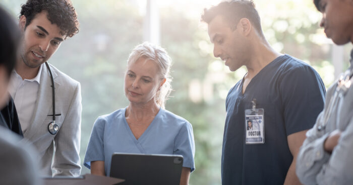 Group of diverse medical professionals in scrubs and a lab coat gathered around a tablet, reviewing patient information together during a hospital team consultation.