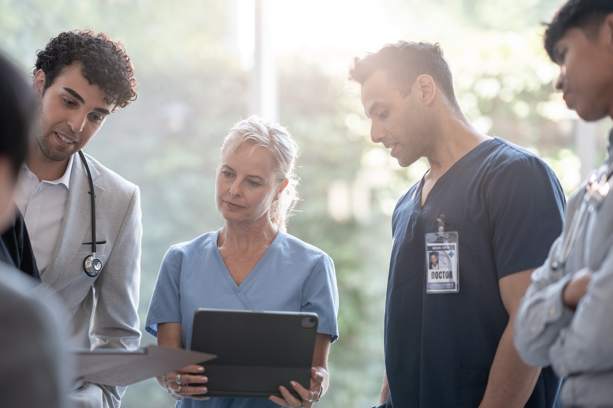 Group of diverse medical professionals in scrubs and a lab coat gathered around a tablet, reviewing patient information together during a hospital team consultation.