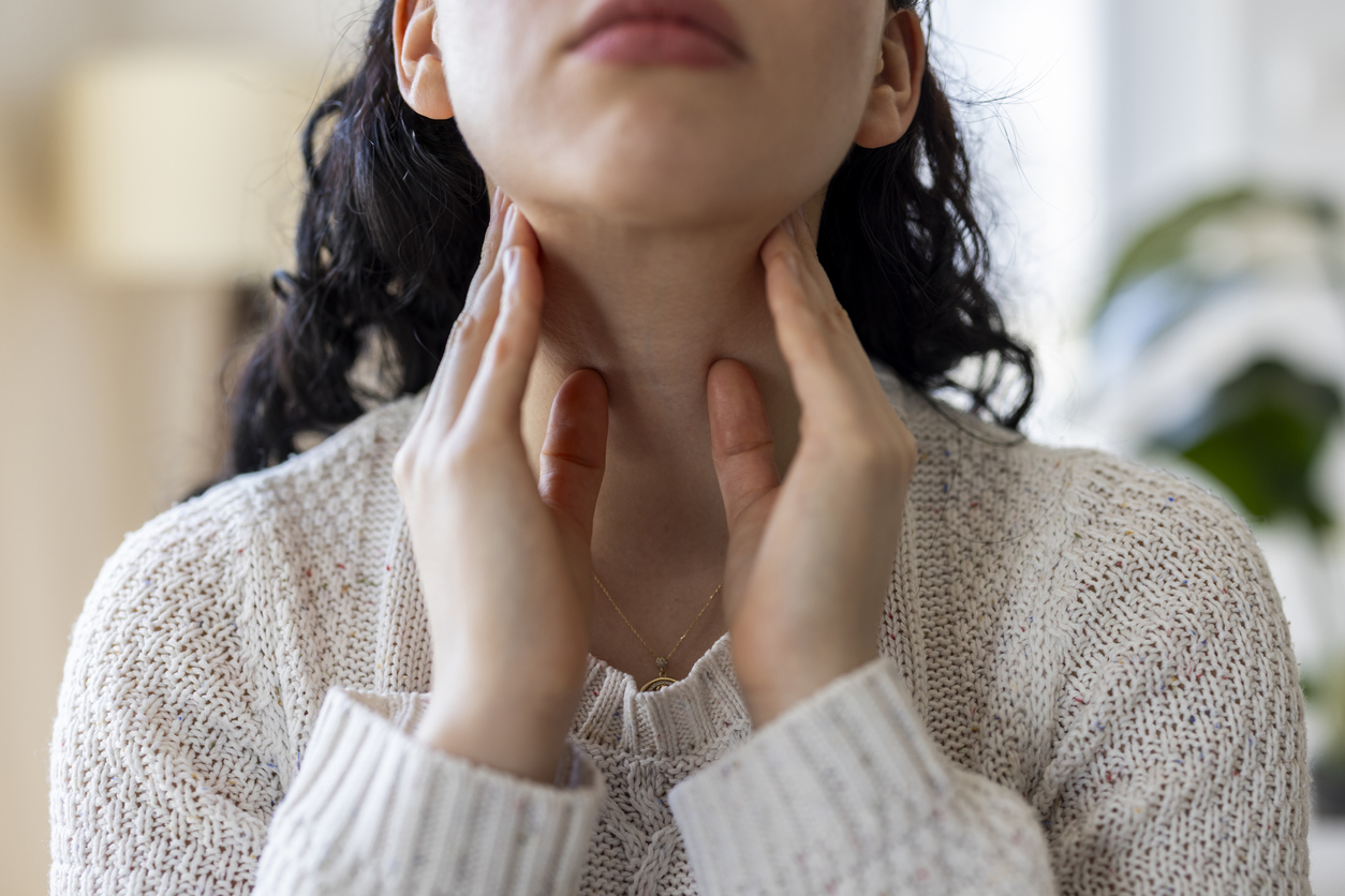 A close-up shot of an unrecognizable young woman touching her neck and throat with her fingers. She is wearing a cozy white knit sweater. The image strongly depicts a physical health condition such as a severe sore throat, swallowing pain, tonsillitis, thyroid gland problems, swollen lymph nodes, or checking for a lump. It represents themes of sickness, medical care, endocrinology, and physical discomfort at home.